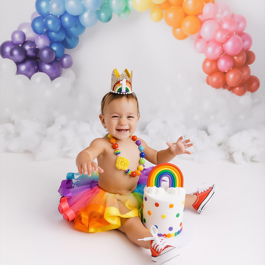 Cake smash photography backdrops - Child in rainbow-themed outfit with balloons and cake, celebrating a birthday.